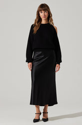 A woman is wearing a black ribbed knit sweater with one shoulder exposed and long sleeves, paired with a long, silky black skirt that reaches her ankles. She is also wearing black high-heeled shoes with ankle straps.